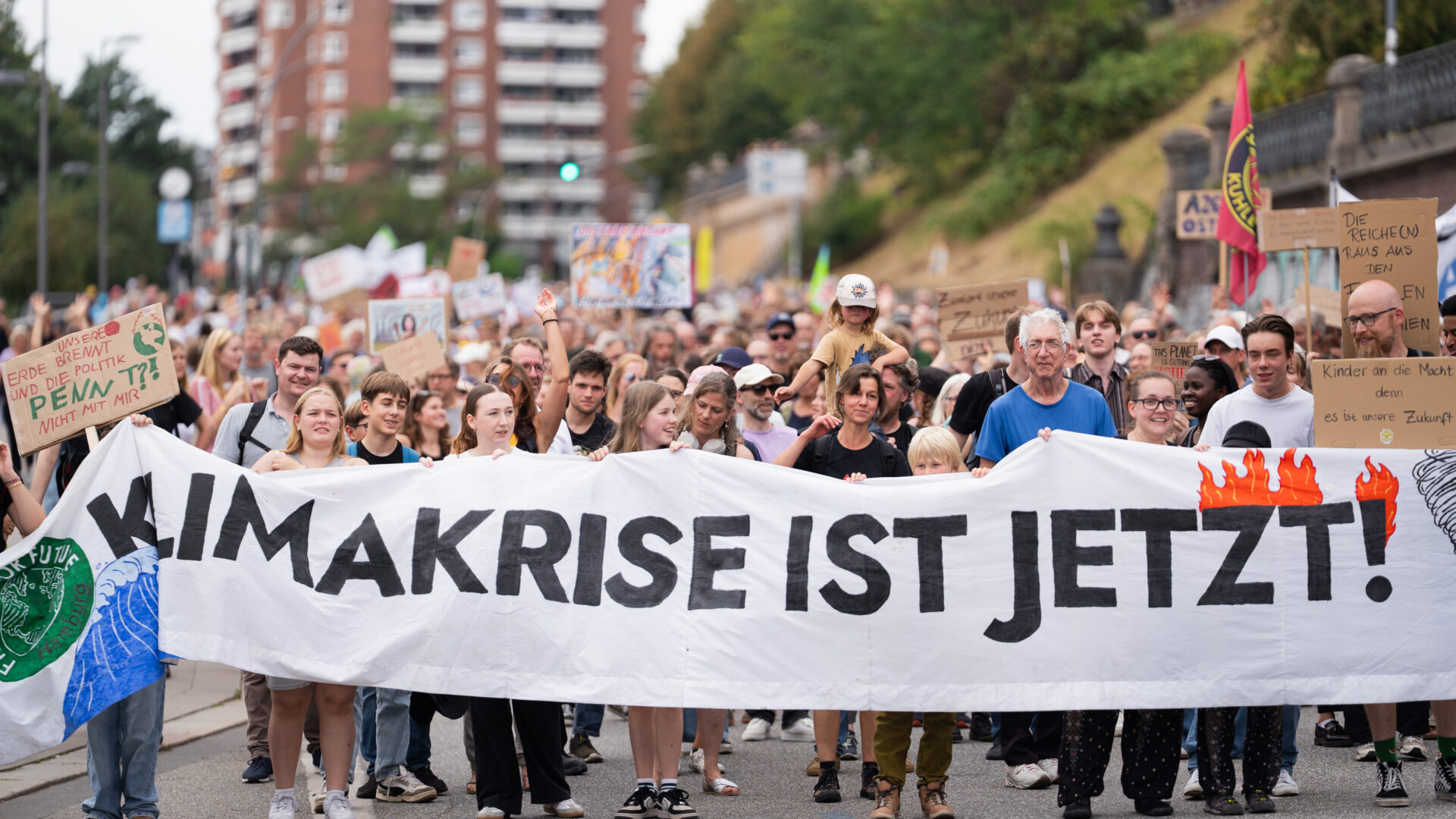 Menschenmenge mit Plakaten auf FFF-Demonstration in Hamburger Innenstadt, Hintergrund verschwommen, im unteren Zentrum des Bildes ein von Demonstrierenden getragenes Banner mit der Aufschrift "Klimakrise ist jetzt!" (ein Teil des Textes in gemalten Flammen)