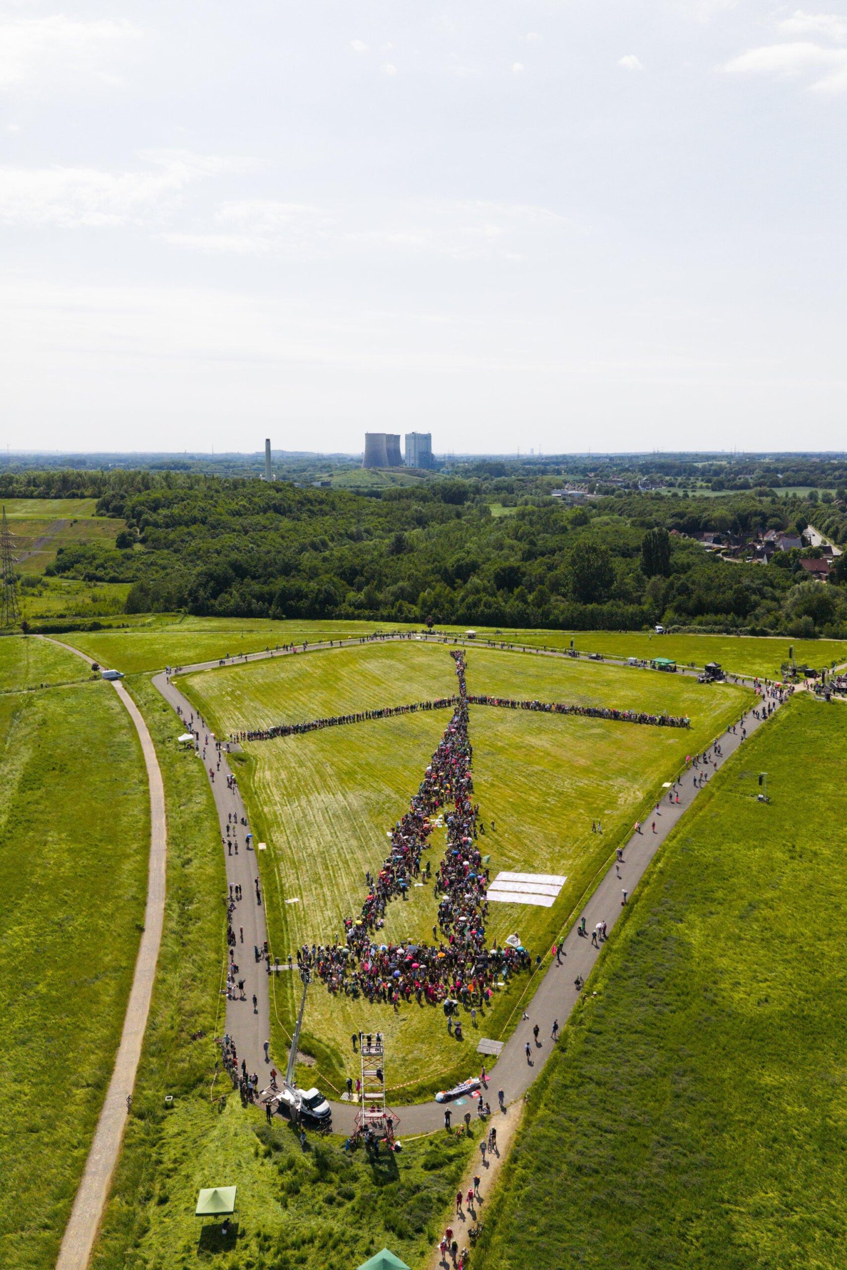 Drohnenbild von einer großen Rasenfläche, darauf ein von mehr als 1000 Menschen gebildetes Windrad (mit Turm und drei Flügeln), im fernen Hintergrund fossile Infrastruktur.