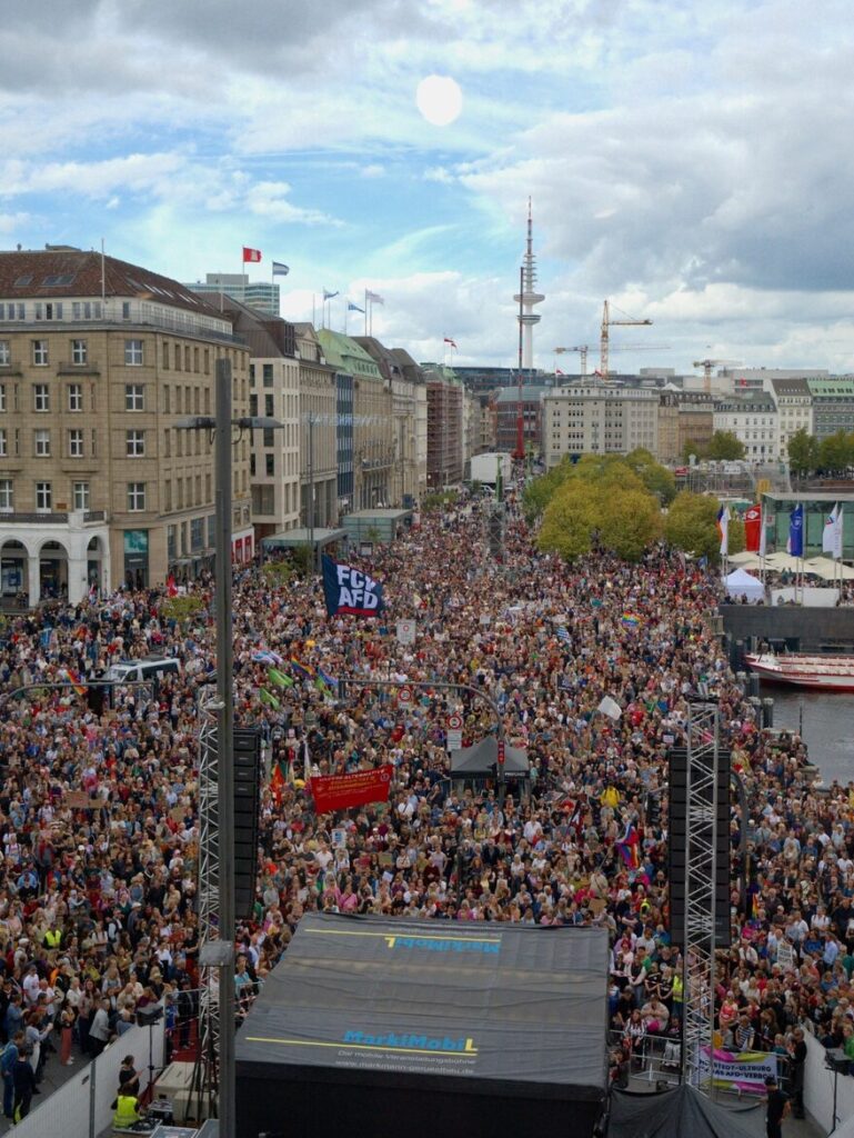 Hintergrundbild: Jungfernstieg, photographiert während der Anfangskundgebung der Hamburger "Zeit zum Handeln"-Demonstration am 12.09.2026, aus recht großer Höhe von hinter der Bühne, komplett menschengefüllt (inklusive Nebenbereiche), Bühne und Backstagebereich teilweise sichtbar, ganz im Hintergrund unter anderem der Gänsemarkt, Fernsehturm und das Radisson.